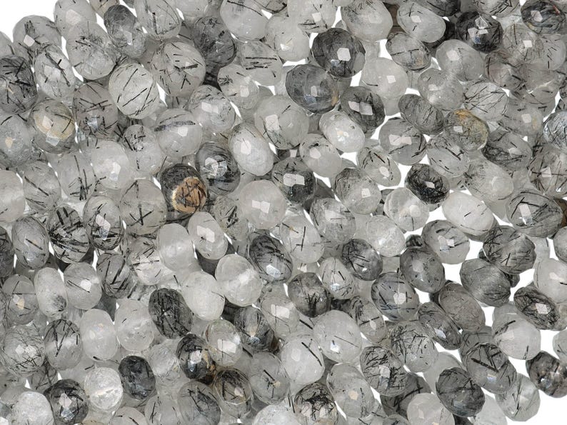 Puede incluir: Un primer plano de una colecci&oacute;n de cuentas de piedras preciosas claras y facetadas con inclusiones negras. Las cuentas de forma ovalada var&iacute;an en tonos de gris y claro, creando una apariencia texturizada. Ideal para la creaci&oacute;n de joyas.