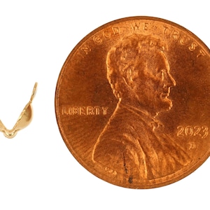 May include: A gold-colored jewelry finding, shaped like a small bra, next to a U.S. penny. The penny shows the profile of Abraham Lincoln and the year 2023. The jewelry finding is likely used in jewelry making.