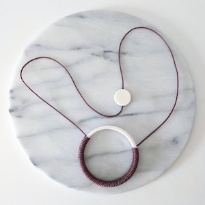 Può includere: Una collana in ceramica bordeaux e bianca con un ciondolo rotondo. La collana è su una superficie di marmo bianco.