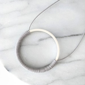 May include: A necklace featuring a large circular pendant with a smooth white upper half and a lower half wrapped in gray cord. The pendant is suspended from a thin gray cord that extends upward from the top of the circular frame.