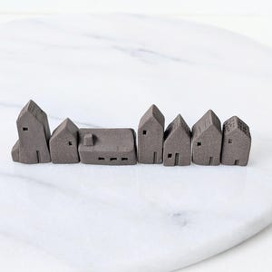 May include: A row of small, gray ceramic houses with varying roof styles and architectural details. Each miniature house features a door and window cutouts, creating a charming village scene. The houses are arranged on a white marble surface.