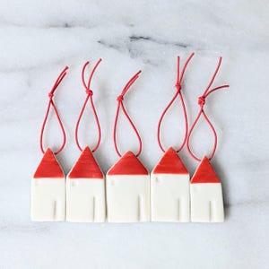 Könnte beinhalten: Fünf Keramik-Hausornamente mit roten Dächern und weißen Körpern, jedes mit einer roten Schnur zum Aufhängen. Die Häuser sind in einer Reihe vor einem Marmorhintergrund angeordnet. Die Ornamente sind ein bezaubernder Dekorationsartikel.