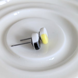 May include: A pair of stud earrings featuring smooth, rounded shapes. One earring is white with a black accent, and the other is white with a yellow accent. Both earrings have silver-toned metal posts and backs, designed for pierced ears.