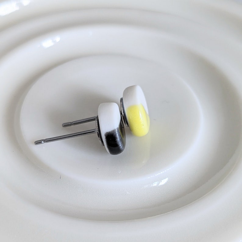 May include: A pair of stud earrings featuring smooth, rounded shapes. One earring is white with a black accent, and the other is white with a yellow accent. Both earrings have silver-toned metal posts and backs, designed for pierced ears.