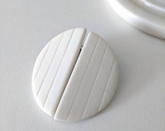 May include: A white ceramic round disc with a grooved design. The disc is split in half and the two halves are slightly offset.