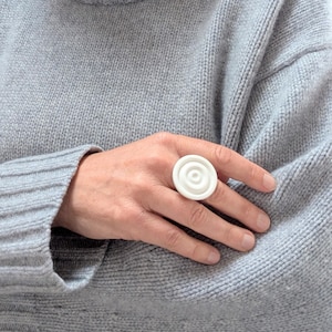 May include: A white ring featuring a circular, multi-layered, concentric design with a raised center point. The ring is worn on a finger, contrasting against the texture of a light gray knit sweater worn in the background of the image.