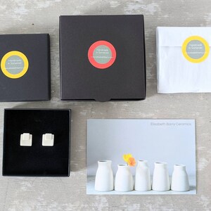 May include: Two black square gift boxes, one white paper pouch, and a pair of small white square ceramic stud earrings inside an open black box. A postcard features five white ceramic vases and the text Elisabeth Barry Ceramics.