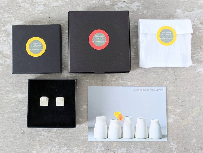 May include: Two black square gift boxes, one white paper pouch, and a pair of small white square ceramic stud earrings inside an open black box. A postcard features five white ceramic vases and the text Elisabeth Barry Ceramics.