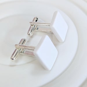 Puede incluir: Un par de gemelos con caras blancas en forma de diamante y varillas de metal plateado. Los gemelos se muestran sobre una superficie blanca.