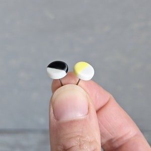 May include: Two small circular stud earrings with flat faces. One earring features a split design of black and white, while the other earring features a split design of pale yellow and white. Each earring has a thin metal post attached to the back.