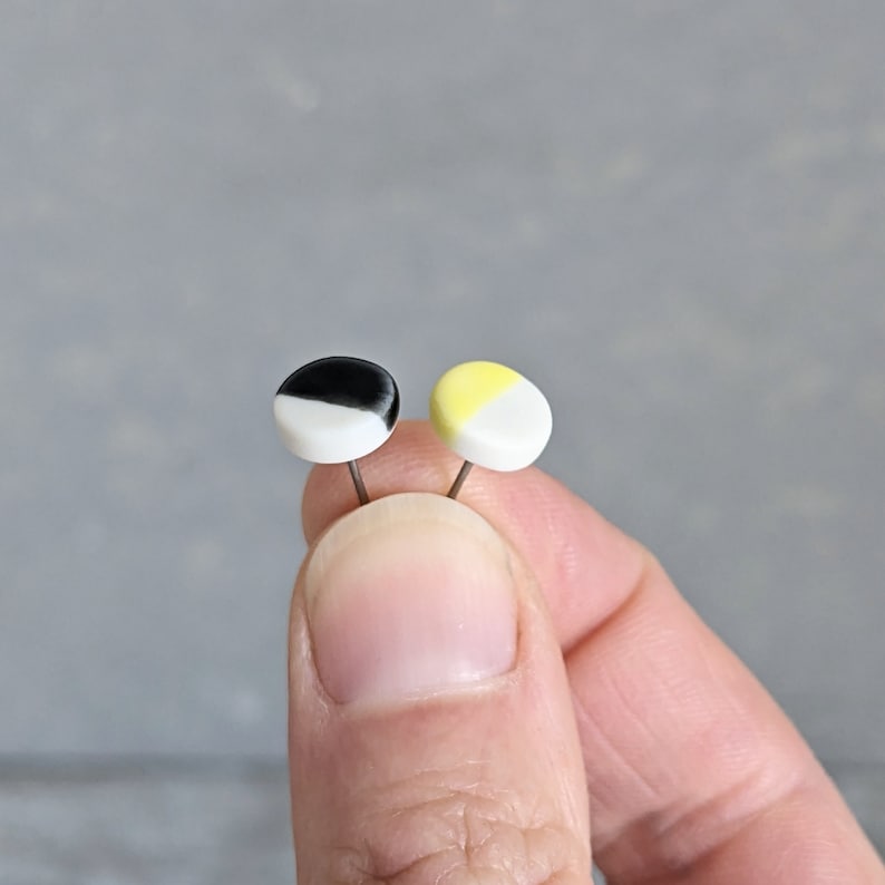 May include: Two small circular stud earrings with flat faces. One earring features a split design of black and white, while the other earring features a split design of pale yellow and white. Each earring has a thin metal post attached to the back.