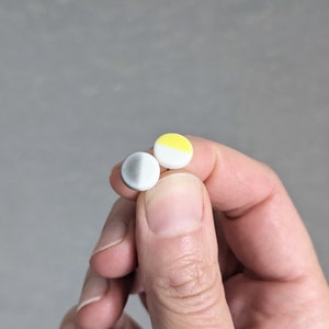 May include: Two small circular stud earrings. One earring features a gradient transition from dark gray to white. The second earring is divided into two distinct halves, with one side colored bright yellow and the other side solid white.