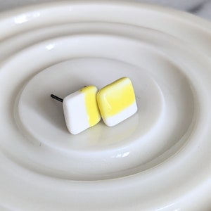 May include: A pair of square stud earrings featuring a smooth, glossy finish. Each earring is divided into two sections, with one half colored in bright yellow and the other half in crisp white. The earrings include dark metal posts for attachment.
