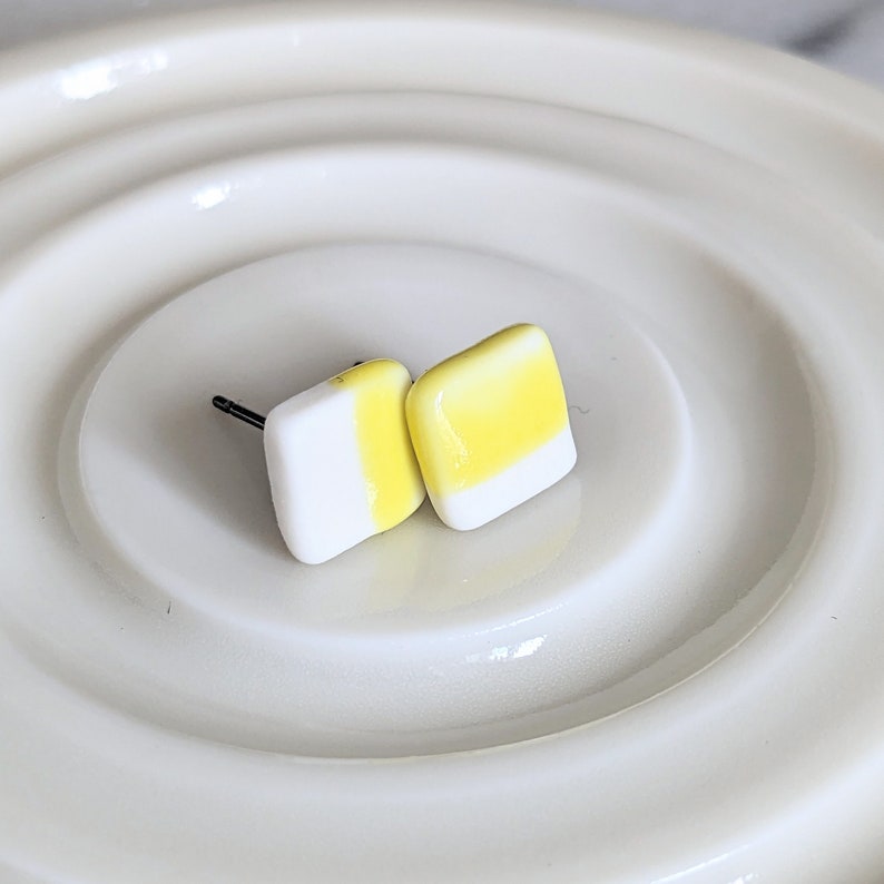 May include: A pair of square stud earrings featuring a smooth, glossy finish. Each earring is divided into two sections, with one half colored in bright yellow and the other half in crisp white. The earrings include dark metal posts for attachment.