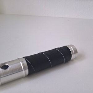 May include: A silver metal tube with a black rubber grip. The tube has a hole in the side and a textured surface.