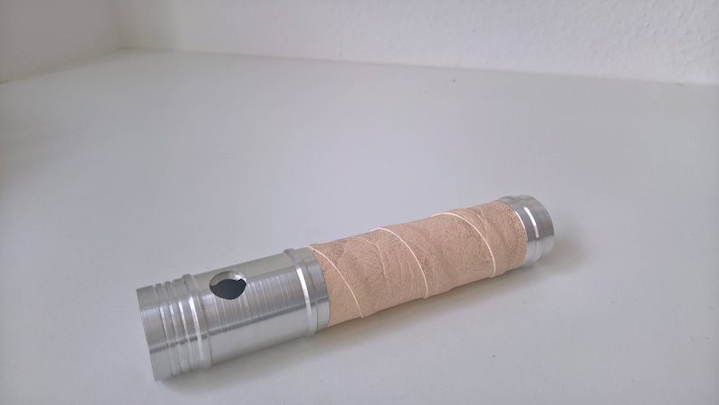May include: A silver metal tube with a brown paper wrap. The tube has a hole in the side and a textured surface.