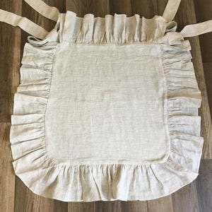 May include: A beige and white striped apron with ruffled edges and long tie straps. The apron is made of a soft fabric and is designed to be worn in the kitchen or for crafting. The apron is laid flat on a wooden surface.