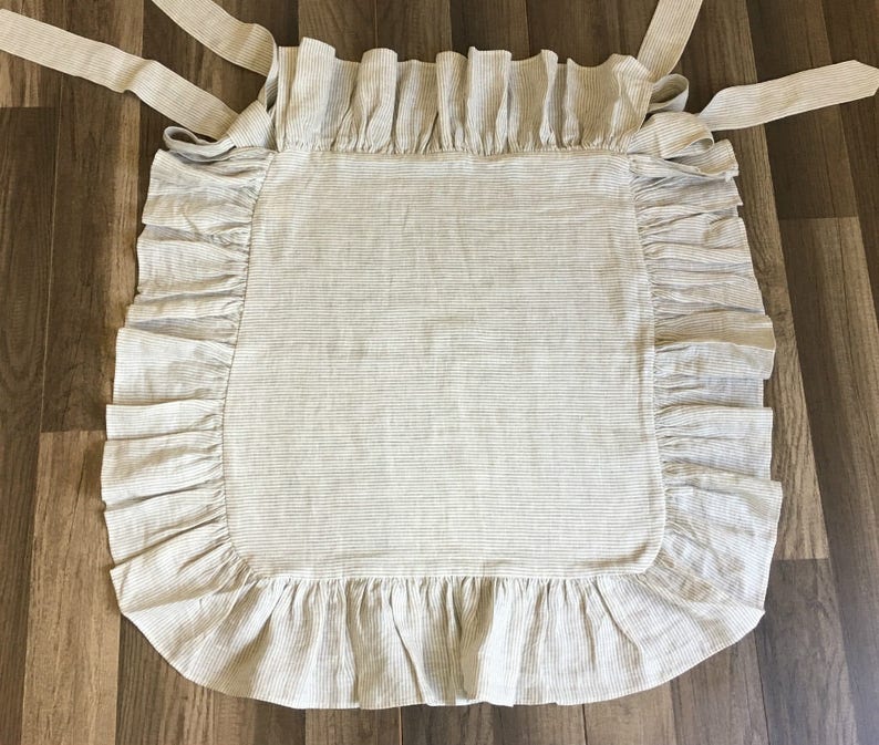 May include: A beige and white striped apron with ruffled edges and long tie straps. The apron is made of a soft fabric and is designed to be worn in the kitchen or for crafting. The apron is laid flat on a wooden surface.