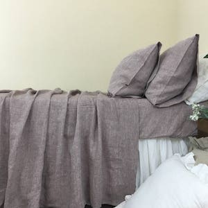 May include: A mauve linen duvet cover with a white linen bed skirt and two mauve linen pillowcases. The bedding is on a bed with a white wall in the background.