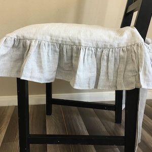 May include: A black wooden stool with a striped cushion cover. The cushion cover is white with thin gray vertical stripes and a ruffled edge. The stool is indoors, with a light-colored wall in the background.