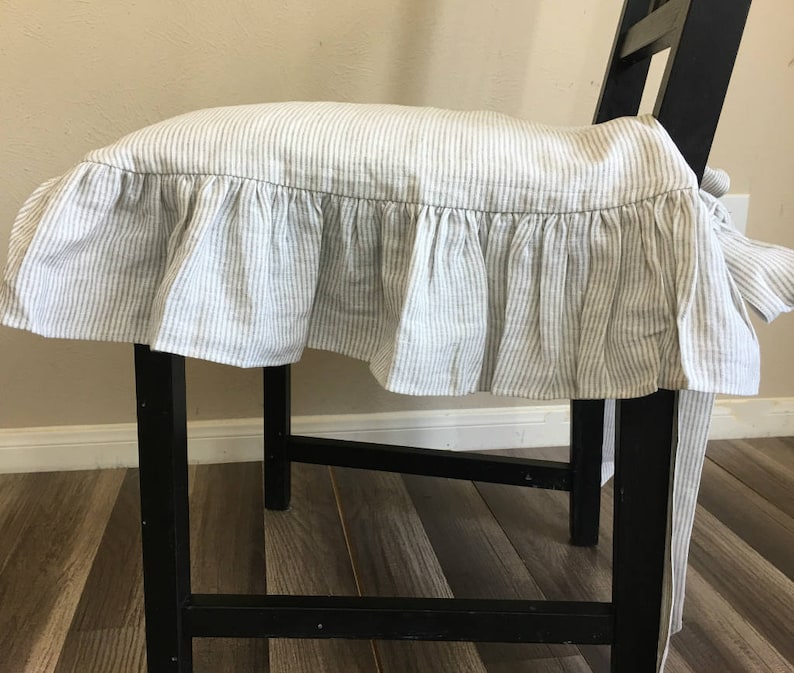 May include: A black wooden stool with a striped cushion cover. The cushion cover is white with thin gray vertical stripes and a ruffled edge. The stool is indoors, with a light-colored wall in the background.