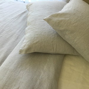 Pu&ograve; includere: Due federe in lino a righe in un'alternanza di colori grigio chiaro e bianco. Le federe sono disposte su un letto con un copripiumino in lino a righe abbinato.