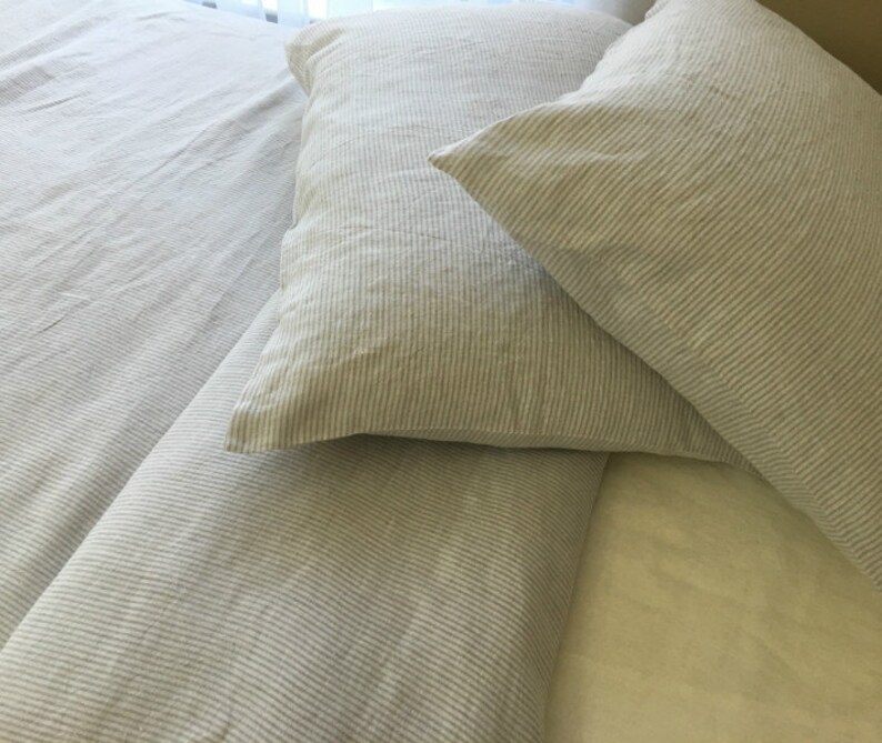 Pu&ograve; includere: Due federe in lino a righe in un'alternanza di colori grigio chiaro e bianco. Le federe sono disposte su un letto con un copripiumino in lino a righe abbinato.