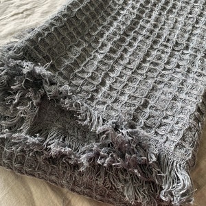 May include: A folded gray waffle-weave blanket with fringed edges. The blanket has a textured, honeycomb-like pattern. The fringe is a lighter shade of gray than the main body of the blanket. The blanket is folded over on itself.