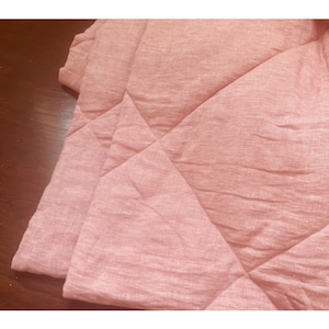 May include: A pink linen quilted throw blanket with a diamond pattern. The blanket is folded in half and the fabric has a subtle texture.