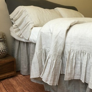 May include: A white linen duvet cover with thin grey stripes and a ruffled edge. The duvet cover is on a bed with a white sheet and two matching pillowcases with ruffles.