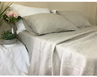 Puede incluir: Una sábana de cama de lino en color beige natural con una textura sutil. La sábana está doblada y se muestra en una cama con dos fundas de almohada de lino a juego.