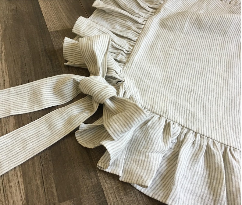 May include: Close-up of a striped apron with a ruffled edge and long tie. The apron is a light gray color with thin white stripes. The tie is knotted in a bow. The apron is made of a soft, textured fabric.