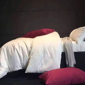 Op de afbeelding: Wit beddengoed met een wit dekbedovertrek, twee witte kussens en een bordeauxrood kussen. Een grijs-witte deken is over het bed gegooid. Een bordeauxrood kussen ligt op de vloer voor het bed.