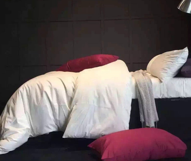 Op de afbeelding: Wit beddengoed met een wit dekbedovertrek, twee witte kussens en een bordeauxrood kussen. Een grijs-witte deken is over het bed gegooid. Een bordeauxrood kussen ligt op de vloer voor het bed.