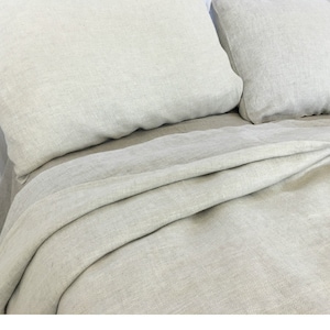 May include: A close-up of a light gray linen bedding set with a herringbone pattern. The bedding is folded neatly on a bed.