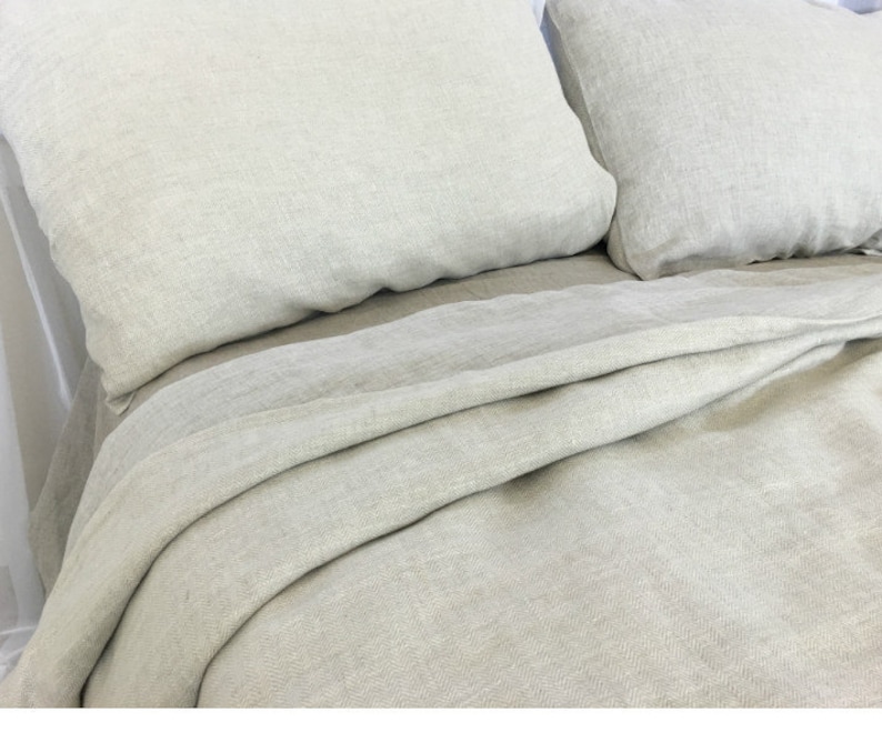 May include: A close-up of a light gray linen bedding set with a herringbone pattern. The bedding is folded neatly on a bed.