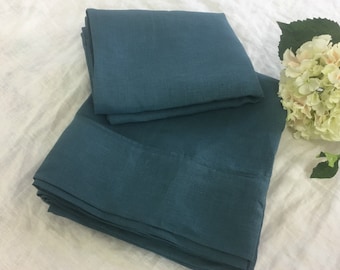 Può includere: Set di biancheria da letto in lino color teal, composto da un lenzuolo piano piegato e un lenzuolo con angoli. Le lenzuola sono realizzate in un tessuto strutturato e sono ben disposte. Un'arrangiamento floreale bianco è sullo sfondo.