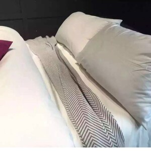 Op de afbeelding: Een wit bed met een wit dekbed en twee witte kussens. Een bordeauxrood kussen ligt aan de linkerkant van het bed. Een grijs-wit gestreepte deken is over het bed gelegd.