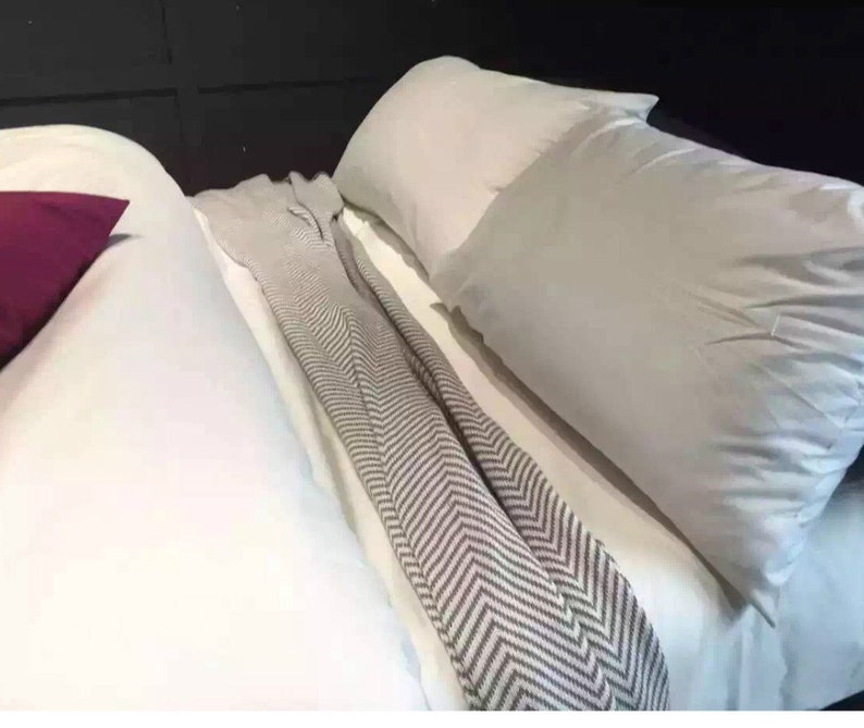 Op de afbeelding: Een wit bed met een wit dekbed en twee witte kussens. Een bordeauxrood kussen ligt aan de linkerkant van het bed. Een grijs-wit gestreepte deken is over het bed gelegd.