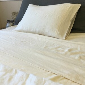 May include: A white linen bed sheet and pillowcase set. The sheet is smooth and wrinkle-free, with a subtle sheen. The pillowcase is folded neatly on top of the sheet.