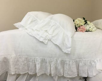 May include: White linen bedding with a ruffled edge and two white ruffled pillowcases. A bouquet of white and pink flowers is placed on the bed.