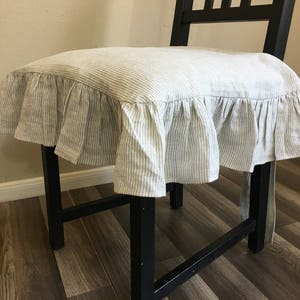 May include: A black wooden chair with a seat cushion cover. The cushion cover is made of a light-colored fabric with vertical stripes and a ruffled edge. The chair is positioned in front of a light-colored wall.