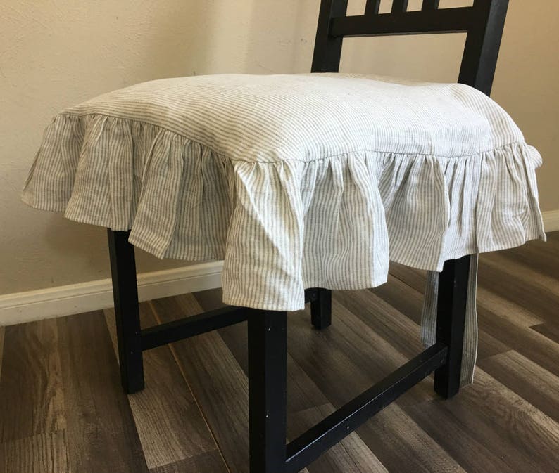 May include: A black wooden chair with a seat cushion cover. The cushion cover is made of a light-colored fabric with vertical stripes and a ruffled edge. The chair is positioned in front of a light-colored wall.