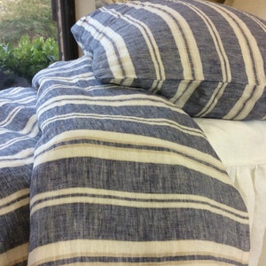 Pu&ograve; includere: Un copripiumino in lino a righe blu e bianche con una riga beige. Il copripiumino &egrave; piegato e posizionato su un letto bianco con un volant bianco.