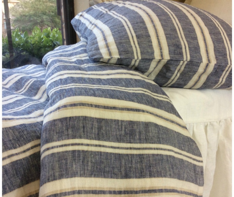 Pu&ograve; includere: Un copripiumino in lino a righe blu e bianche con una riga beige. Il copripiumino &egrave; piegato e posizionato su un letto bianco con un volant bianco.