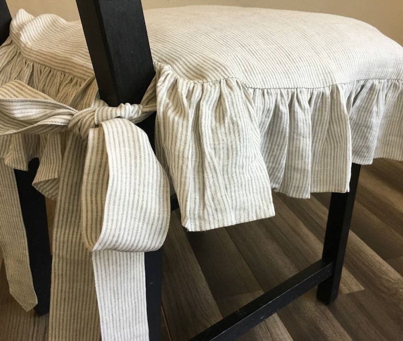 May include: Close-up of a chair with a striped, ruffled seat cover. The cover is tied to the chair legs with a large bow. The fabric has a neutral color palette with vertical stripes.