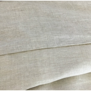 May include: Two pieces of beige linen fabric, one with a herringbone pattern and the other with a plain weave. The fabric is folded and the edges are visible.