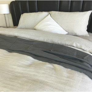 May include: A bed with a gray linen throw blanket on top of a white linen duvet cover. The bed has a black leather headboard and two white linen pillows.