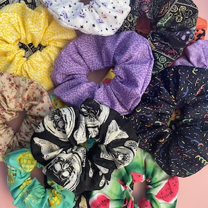 May include: A collection of colorful fabric scrunchies with various patterns, including floral, geometric, and cartoon characters. The scrunchies are made of soft, stretchy fabric and are perfect for holding hair in place.
