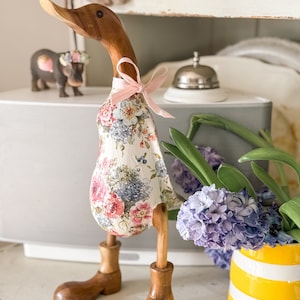 May include: A decorative wooden duck figurine, featuring a floral dress and brown boots. The duck has a wooden head and legs, and a pink ribbon tied around its neck. The dress displays pink, blue, and white flowers. A charming home decor item.